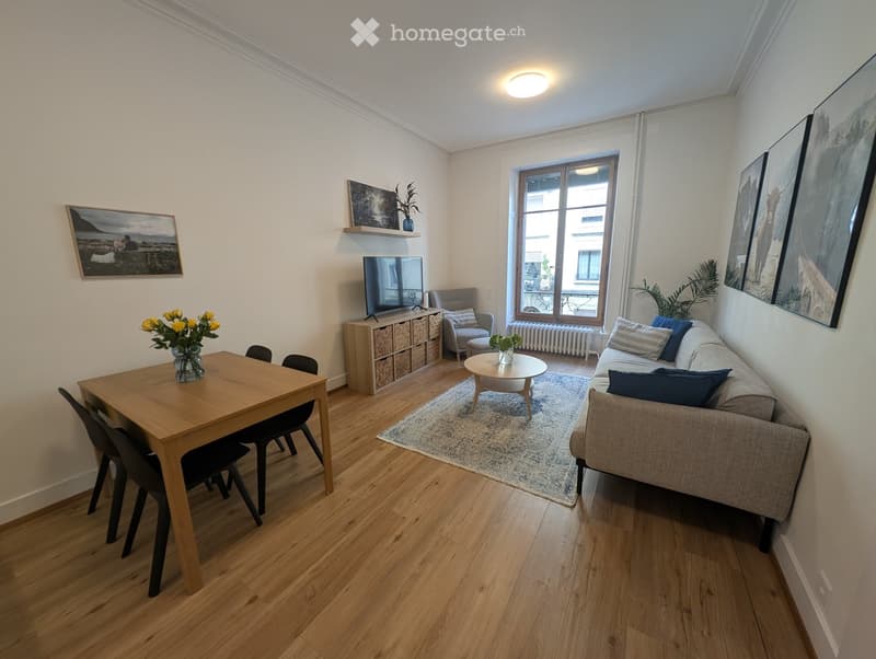 Bright living room with wooden flooring, a sofa, coffee table, dining table with chairs, TV, storage unit, and decorative artwork.
