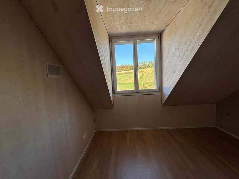 Empty attic room with wooden flooring, slanted wooden ceiling, and a large window offering a view of a green field.