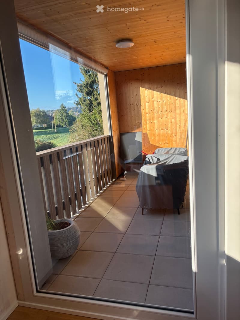 Balcony with wooden railing, tiled floor, covered table and chairs, potted plant, and scenic view of greenery.