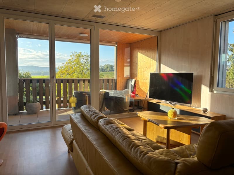 Modern living room with wooden floors, large windows, and a view of the countryside. Features a beige leather sofa, wooden coffee table, flat-screen TV, and a small table with a decorative item.