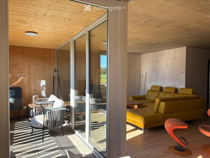 Modern living room with wooden walls and ceiling, large glass sliding doors leading to a patio, a yellow leather sofa, a coffee table, a wicker chair, and colorful stools.