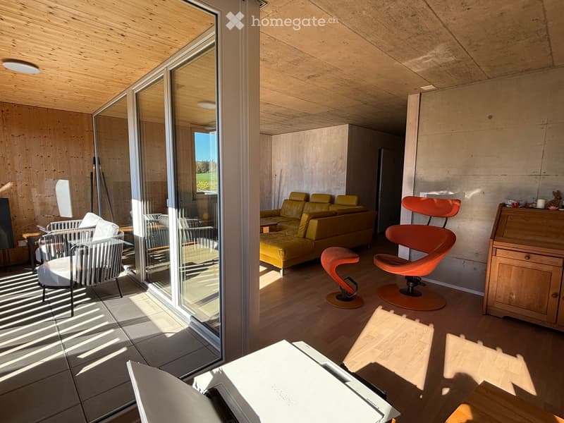 Modern living room with wooden floors, large glass doors, a yellow sofa, orange chairs, and a wooden table with a laptop.