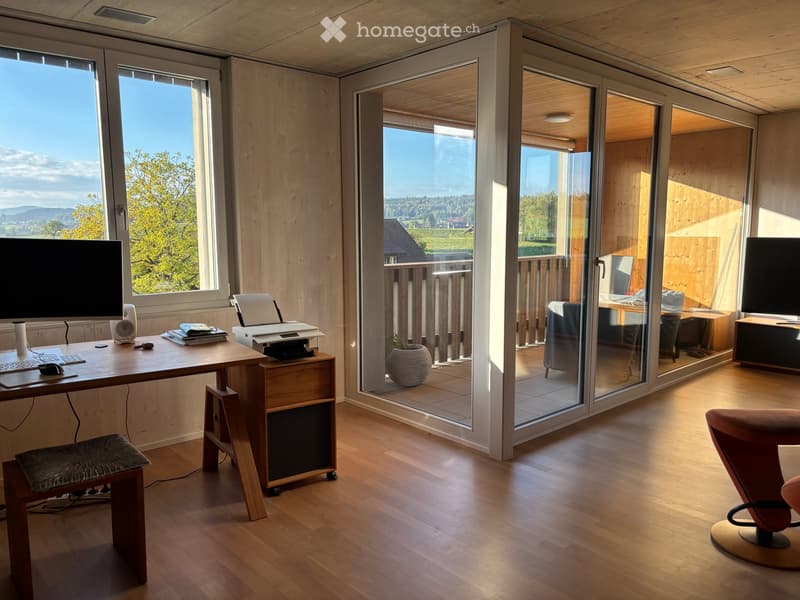 Bright, modern room with wooden flooring and large windows offering scenic views. Features a desk with a computer setup, a comfortable chair, and a cozy seating area with a red armchair.