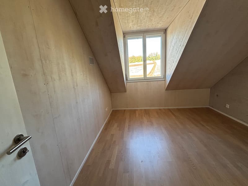 Empty attic room with wooden flooring, slanted ceilings, and a window offering a view of the outdoors.