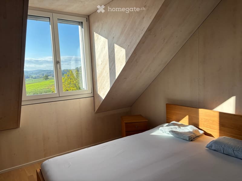 Cozy attic bedroom with wooden walls and slanted ceilings, featuring a large window with scenic countryside view, a neatly made bed with white linens, a nightstand, and a folded blanket on the bed.