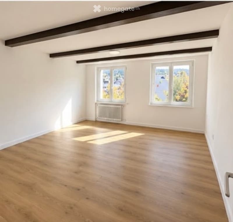 Empty room with wooden flooring, white walls, and two windows offering a view. Exposed wooden beams on the ceiling.