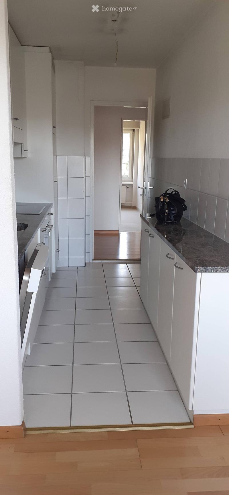 A clean, modern hallway with white tiled walls, wooden flooring, and a view into a kitchen area with white cabinets and a granite countertop.