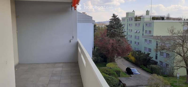 Balcony with tiled floor, white railing, and view of surrounding buildings, trees, and parked car.
