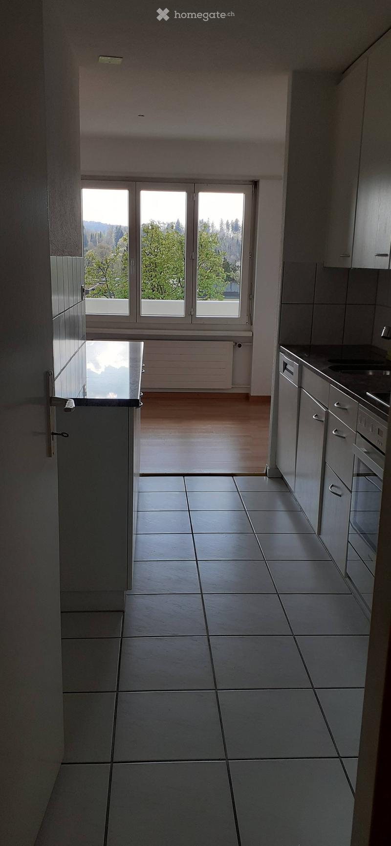 A modern, minimalist hallway with tiled flooring, white walls, and a large window at the end leading to a balcony area with wooden flooring and a view of greenery.