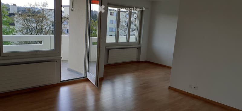 Empty room with wooden flooring, large windows, and a balcony view of trees and buildings.