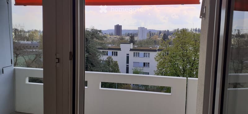 Balcony with white railing, overlooking a cityscape with trees, buildings, and distant hills.