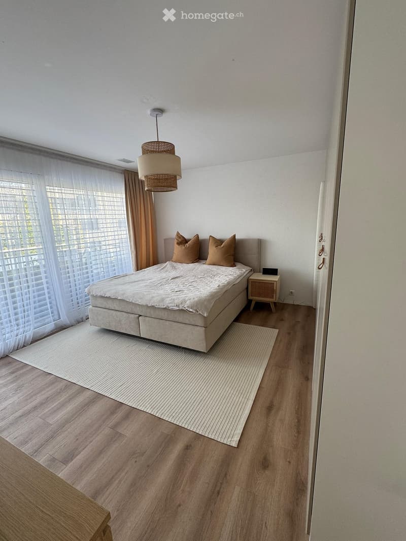 Modern bedroom with a large bed, beige bedding, two throw pillows, a pendant light fixture, a nightstand, and a white rug on a wooden floor.