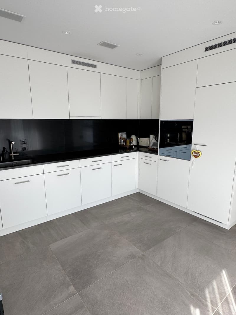 Modern kitchen with white cabinets, grey tiled floor, and stainless steel appliances including a sink, microwave, and toaster. The countertops are black and grey.