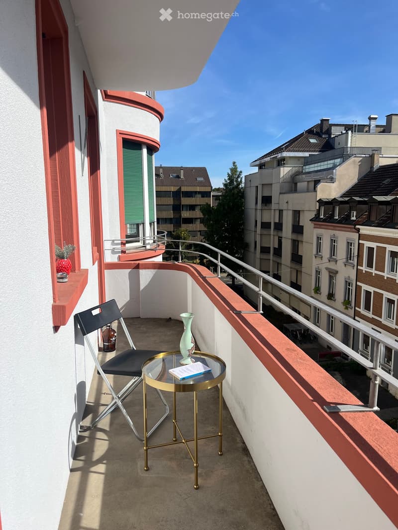 Balcony with a small round table, a chair, a vase, and a book, overlooking a cityscape with buildings and trees.
