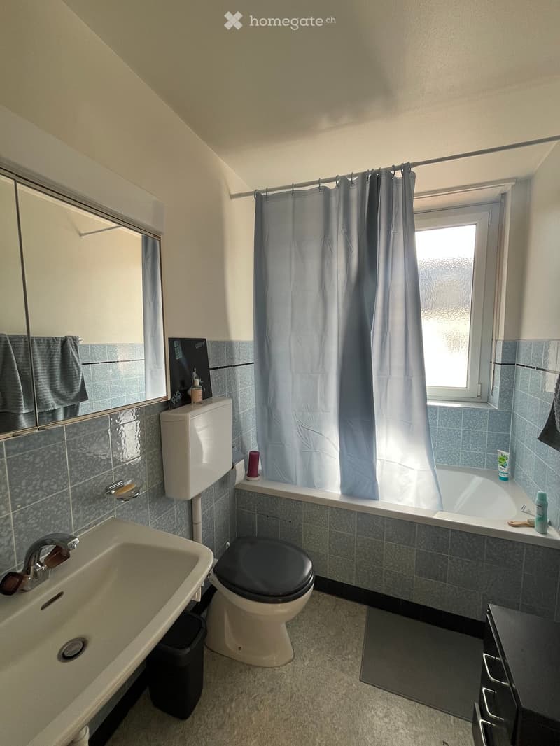 Bathroom with a toilet, sink, bathtub, mirror, and shower curtain. The room features tiled walls and a window above the bathtub.