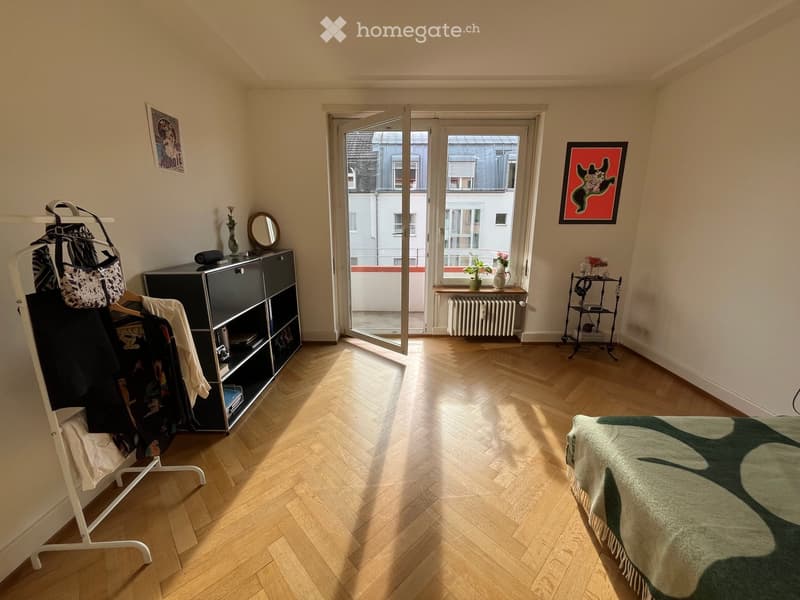 Bright bedroom with wooden herringbone flooring, large window with open door leading to balcony, bed with green and white patterned bedspread, wardrobe, mirror, small table with vase and picture frame, radiator, and clothing rack.