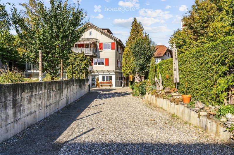 A two-story house with a balcony and red shutters, surrounded by greenery and a gravel driveway.