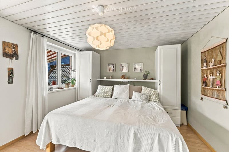 Cozy bedroom with a white bed, patterned pillows, wooden floor, large window with a view, shelves with decorative items, and a hanging light fixture.