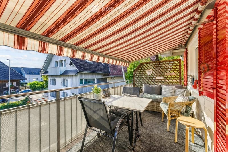 Balcony with a striped awning, a small seating area including a sofa, chairs, a table, and a stool, with a view of neighboring houses.