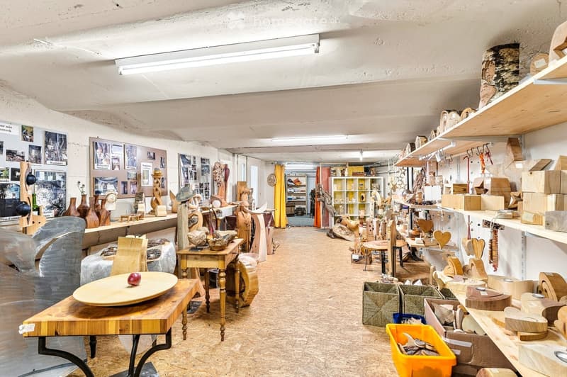 A spacious, well-lit studio or workshop with wooden floors, exposed white walls, and various wooden sculptures and decorative items displayed on tables, shelves, and the floor. The room features a mix of functional furniture and artistic displays, includi