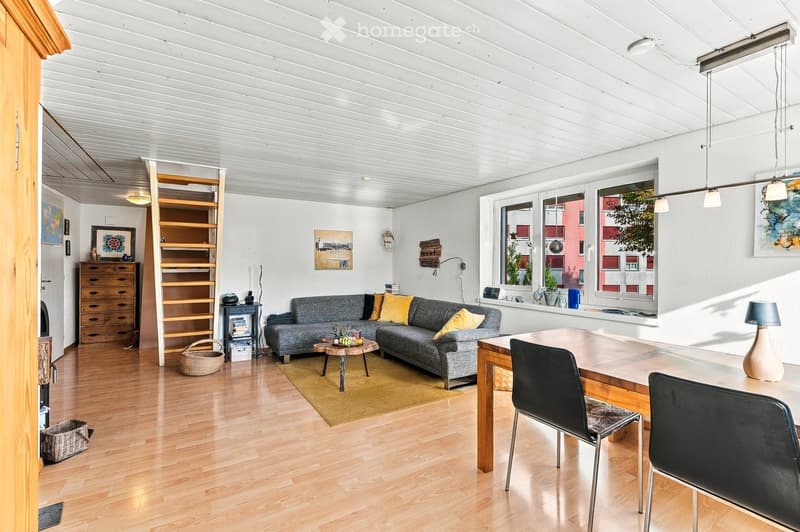 Bright living room with wooden flooring, grey sofa with yellow cushions, coffee table, dining table with chairs, shelves, and decorative items.