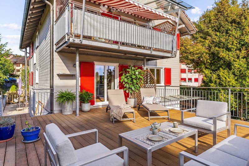 A wooden deck terrace with lounge chairs, a coffee table, and potted plants, featuring a striped awning and a view of the surrounding greenery.