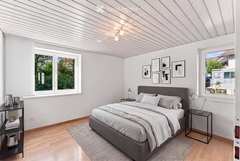Modern bedroom with a large bed, nightstands, lamps, and artwork on the walls. The room features wooden flooring and large windows with a view of greenery.