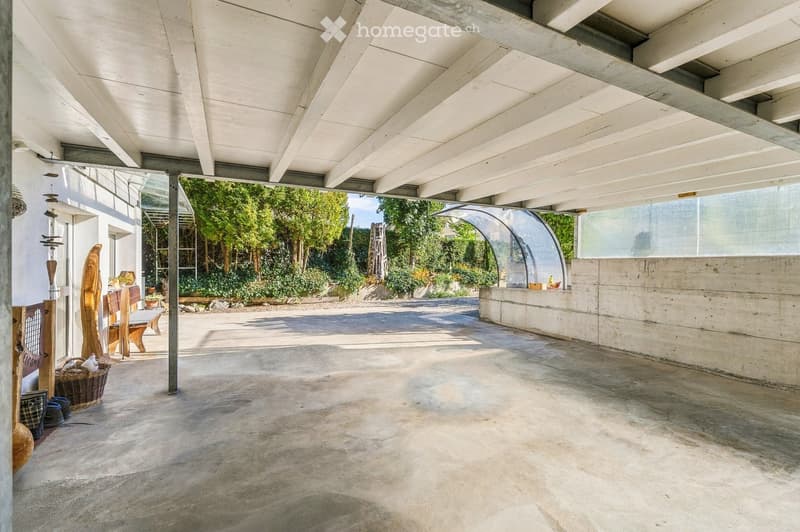An open-air patio with a concrete floor, white ceiling beams, and a view of greenery and trees in the background. There is a small seating area with a bench and a few decorative items, including a wicker basket and a wooden sculpture.