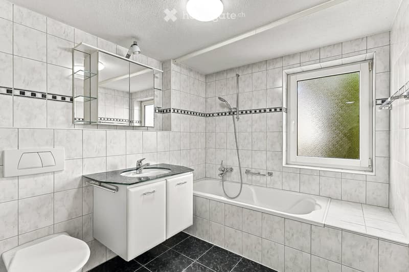 Modern bathroom with white tiles, a large mirror, a sink with a glass countertop, a bathtub, and a toilet. The space features a frosted window and built-in storage cabinets.
