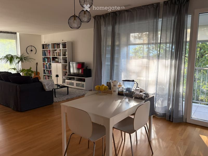 Bright living and dining area with wooden flooring, large windows, and a balcony access. Features a sofa, dining table with chairs, bookshelf, television, and decorative items.