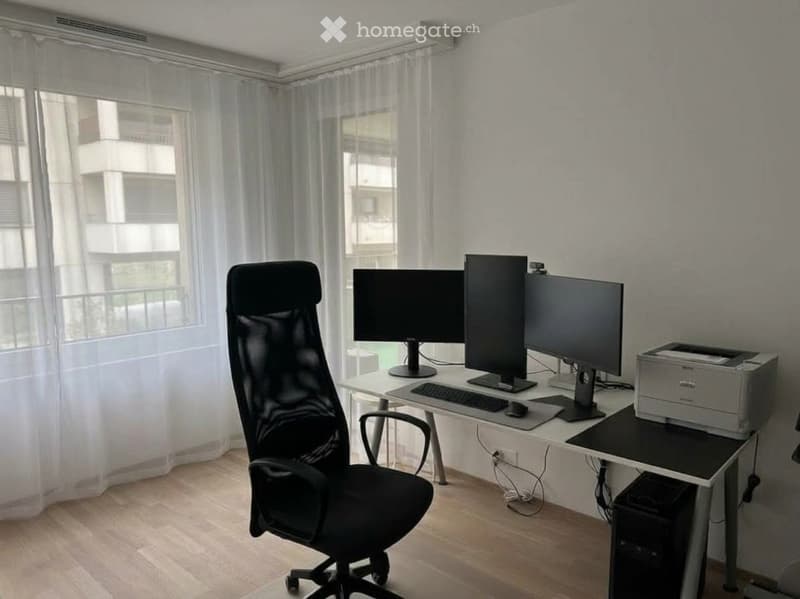 A modern office space with a large desk, multiple computer monitors, a comfortable office chair, and a printer. The room features large windows with sheer curtains and wooden flooring.