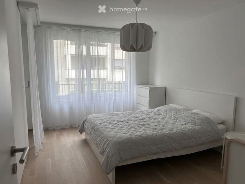 Minimalist bedroom with a white bed, grey bedding, a nightstand, and a modern hanging lamp. The room features light wooden flooring and large windows with sheer curtains.