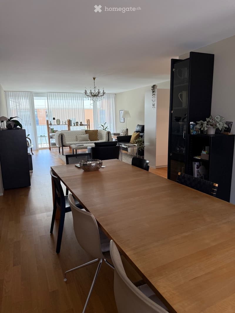 Modern dining and living area with wooden flooring, a long dining table with chairs, a sofa, armchairs, a bookshelf, and a chandelier.