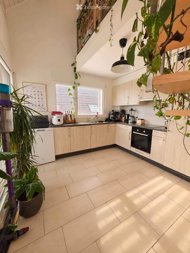 Modern kitchen with light wooden cabinets, tiled floor, and various appliances including an oven, coffee maker, and microwave. There is a large window with a view of a tiled roof, and several plants are placed around the room.