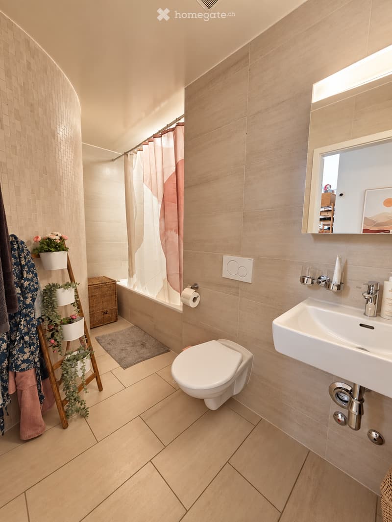 Modern bathroom with light wood tiles, a white toilet, sink, bathtub, and a shower area with a pink curtain. Includes a mirror, towel rack, and decorative plants.