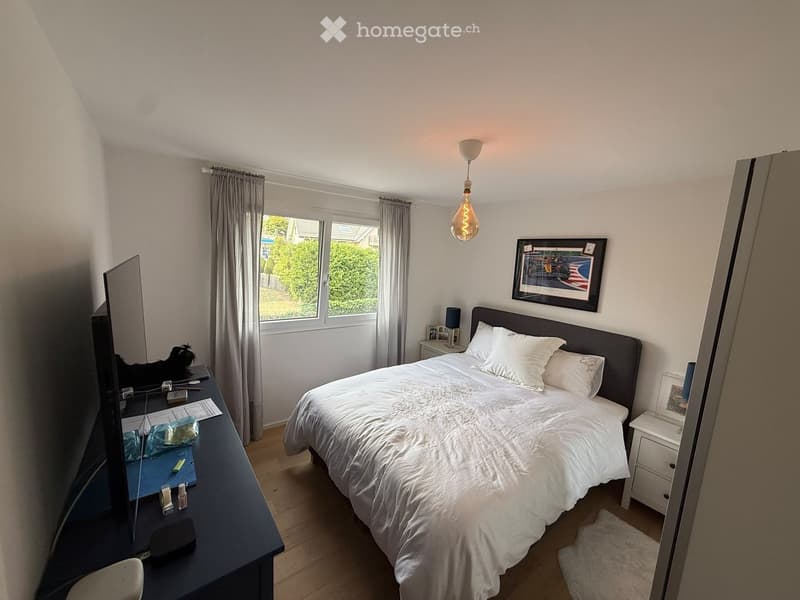 A well-lit bedroom with a large bed, nightstands, a mirror, a chair, and a table with various items on it. The room features wooden flooring and large windows with curtains.