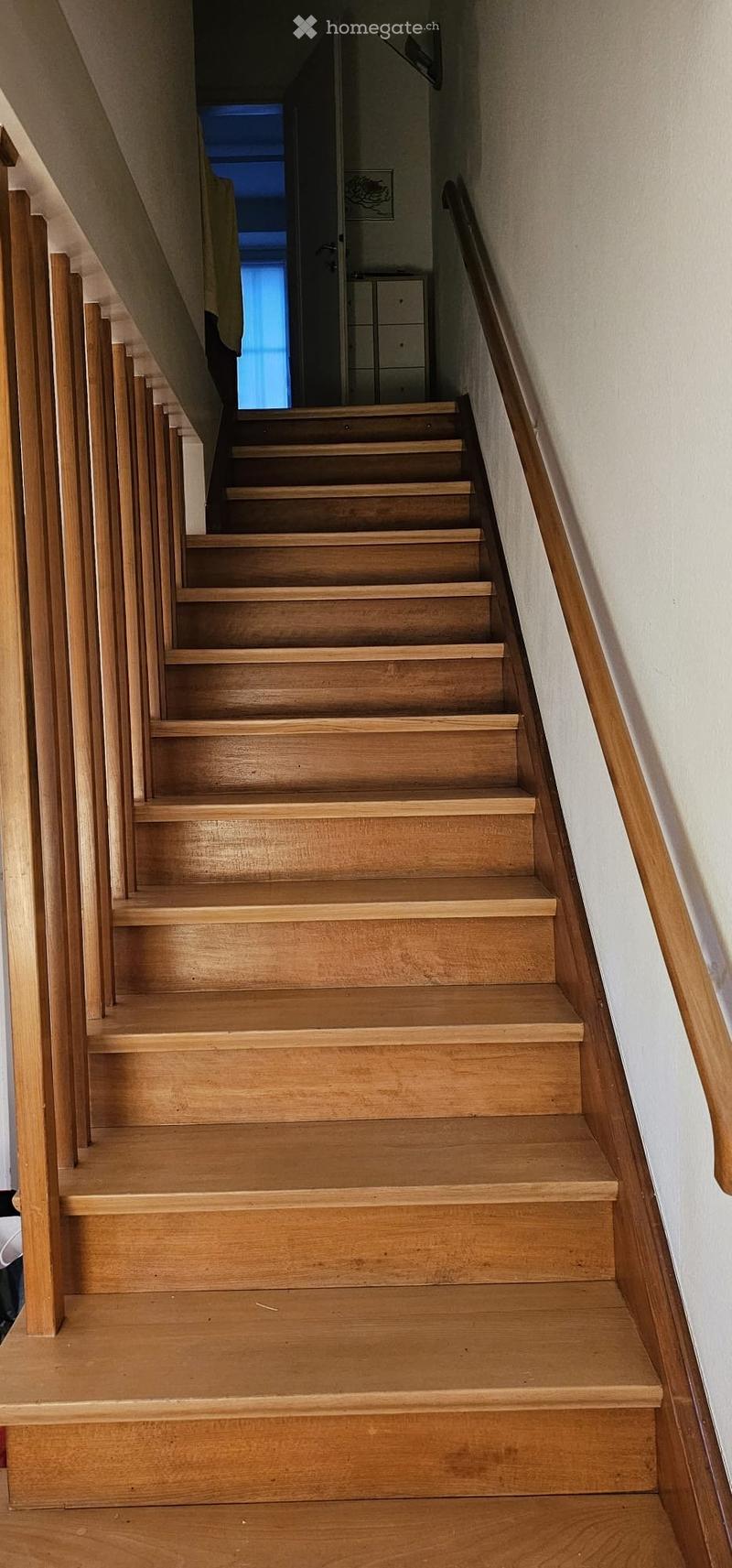 Wooden staircase with a handrail, leading to an open area with a visible door and window at the top.