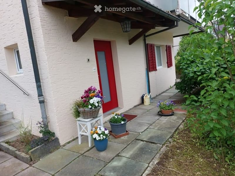 Front entrance of a house with a small garden area, featuring a red front door, a white bench with a flower basket, potted plants, and a small pathway leading to the door.