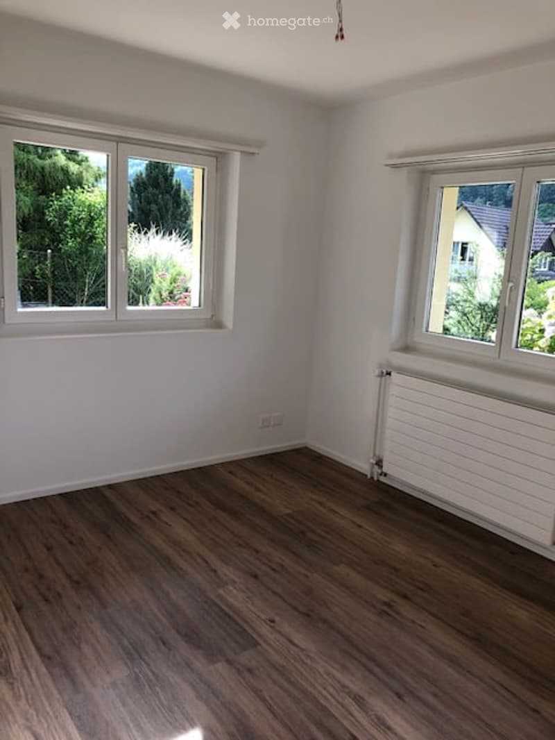 Empty room with wooden flooring, white walls, and large windows offering a view of greenery outside.