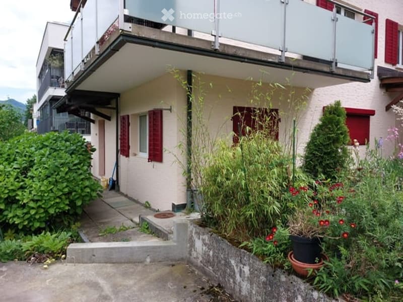 Two-story residential building with a balcony, red shutters, and a small garden area with potted plants and shrubs.