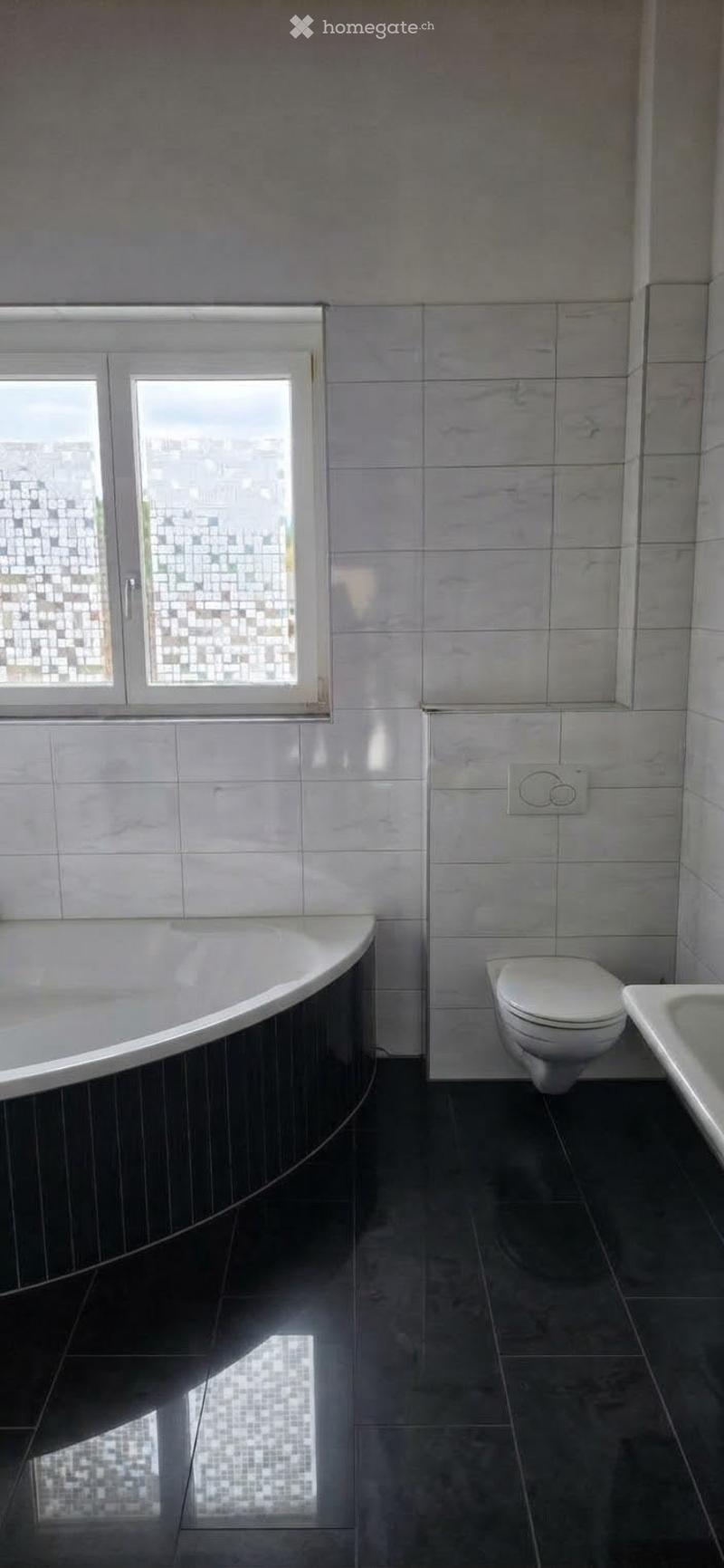 Modern bathroom with a bathtub, toilet, and sink. The walls and floor are tiled in a light grey marble pattern, and there is a window with frosted glass panes.