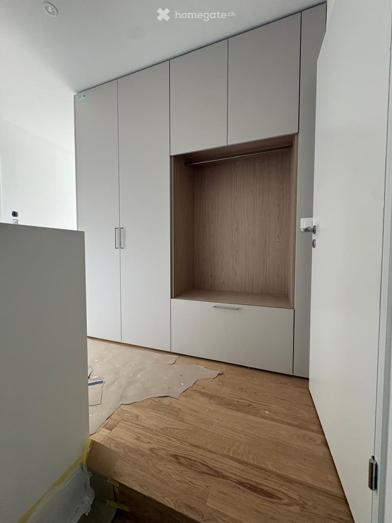 Empty room with wooden flooring and white built-in wardrobes, featuring a partially open closet with wooden shelves.