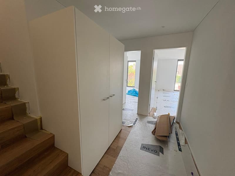 Empty hallway with wooden flooring, white walls, and a staircase leading upwards. The area appears to be under renovation with protective coverings on the floor and some scattered items.
