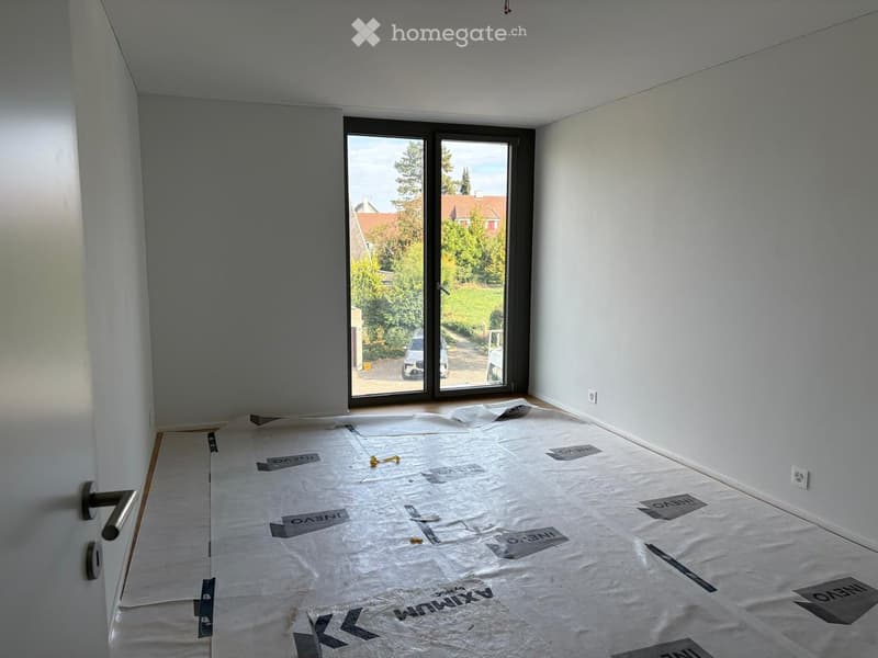 Empty room with white walls, wooden floor covered with protective plastic sheet, large sliding glass door leading to outdoor area.