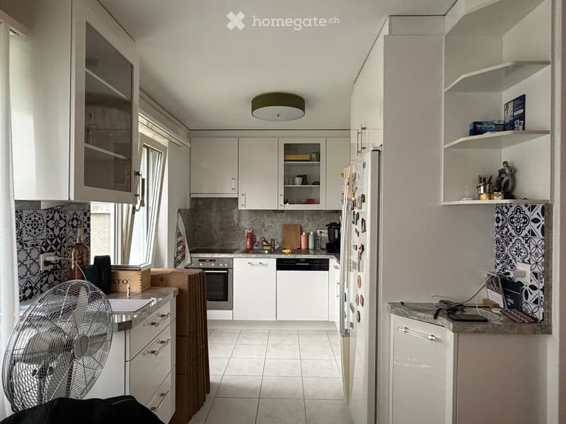 Modern kitchen with white cabinets, tiled backsplash, and stainless steel appliances including a refrigerator, oven, and dishwasher. Countertops hold a fan, various kitchen utensils, and small items.