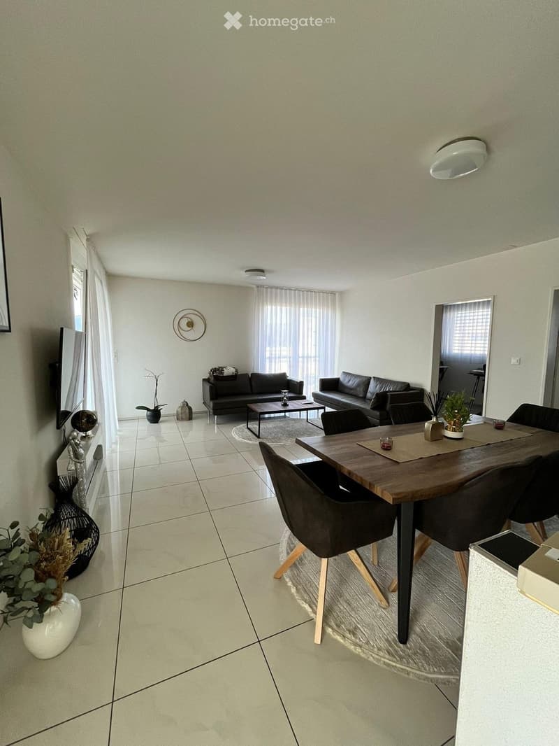 Modern living and dining area with tiled flooring, large windows with curtains, a black leather sofa, a wooden coffee table, a dining table with chairs, decorative vases, and a wall-mounted mirror.