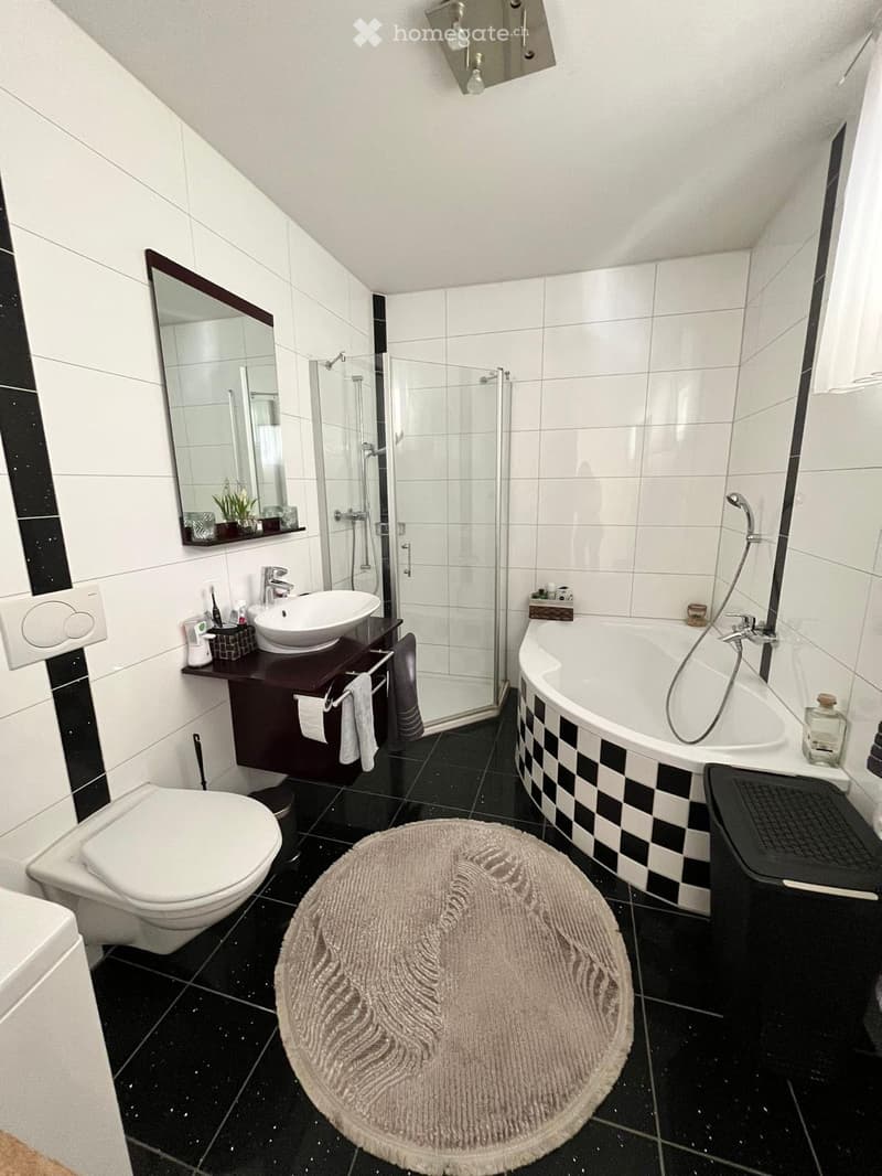 Modern bathroom with black and white tiles, a bathtub with a checkered border, a toilet, a sink with a dark wooden cabinet, a large mirror, and a round rug on the floor.