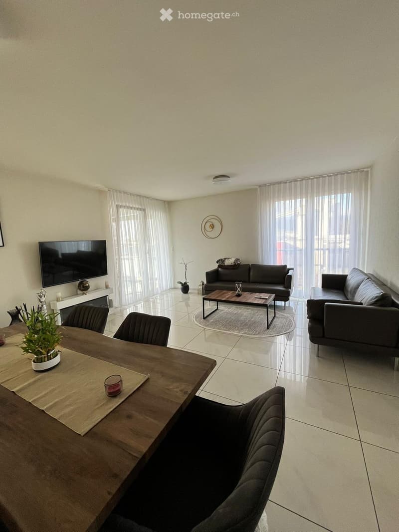 Modern living room with tiled flooring, large windows with sheer curtains, a dark grey sofa, a coffee table, a dining table with chairs, and a television mounted on the wall.