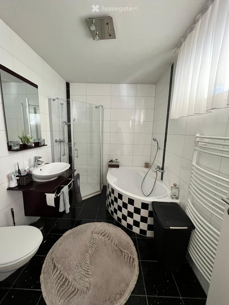 Modern bathroom with black and white checkered bathtub, white sink, toilet, and a large mirror. Features a glass-enclosed shower, towel rack, and a plush round rug on the floor.