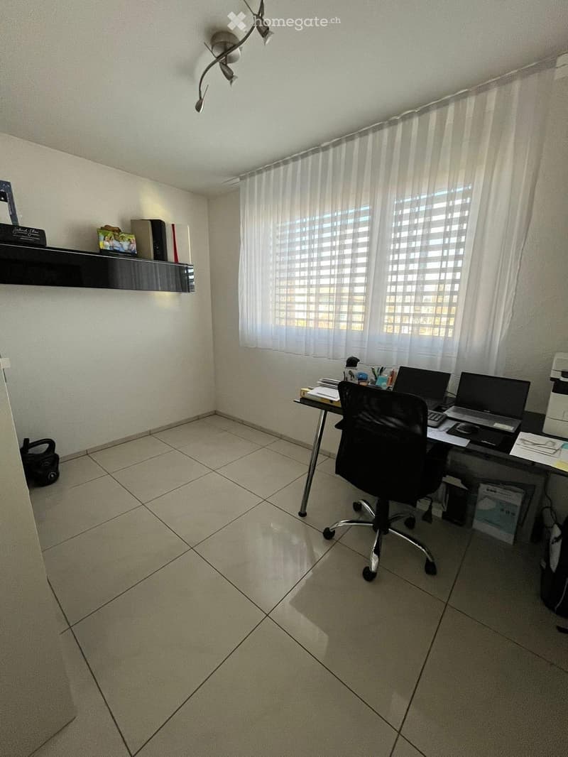 Office space with tiled floor, large window with sheer curtains, desk with chair, laptop, printer, and shelf with books and picture frame.